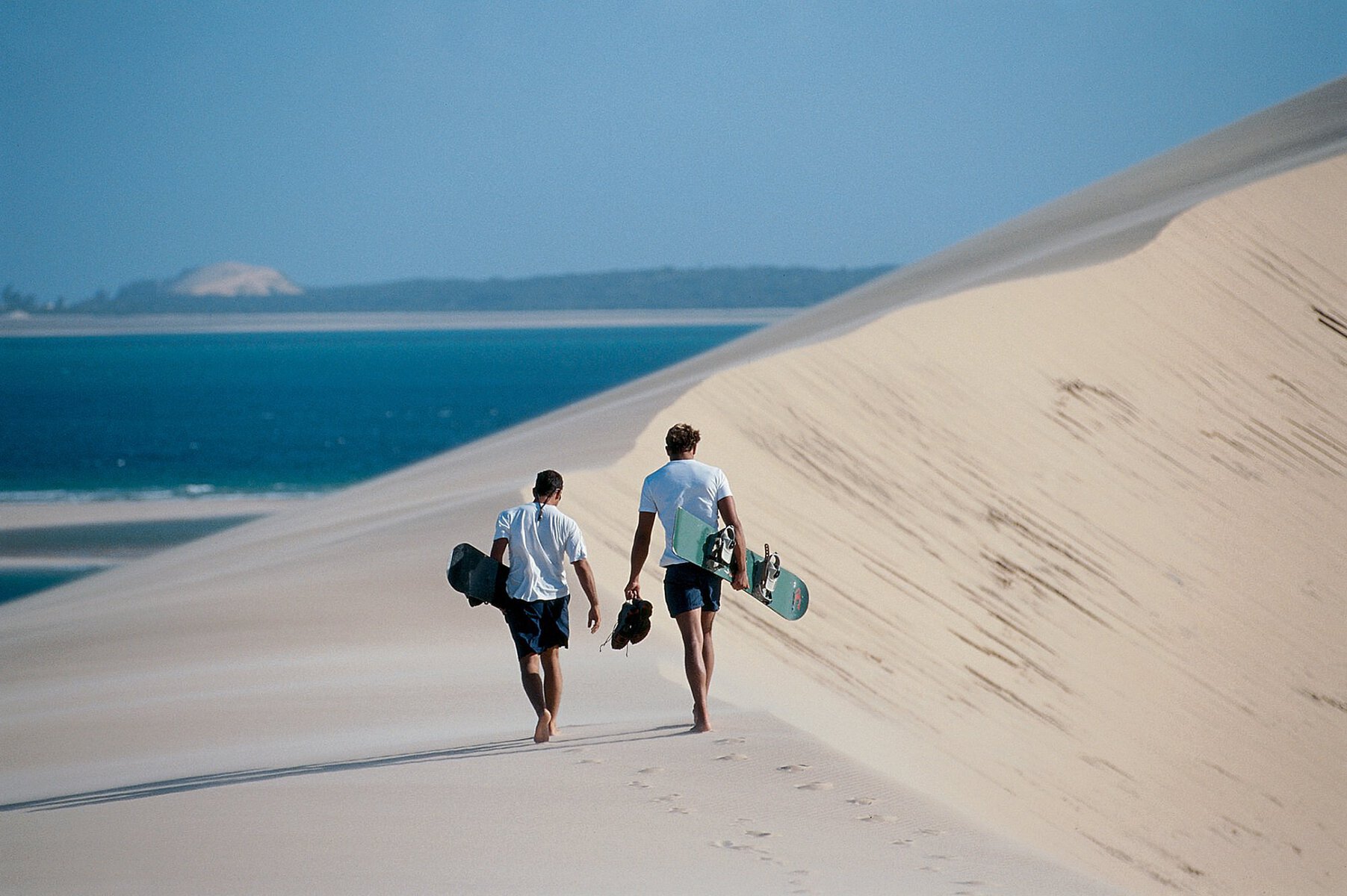 Two people holding sandboards while walking on a sand dune in Mozambique.