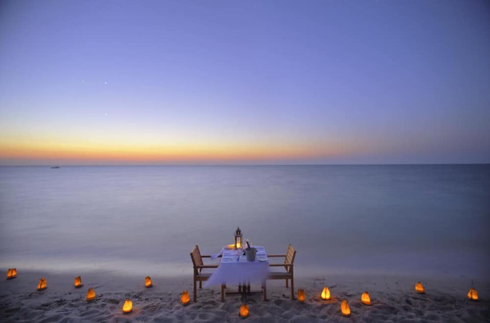 Lanterns on a beach with table and two chairs at sunset.
