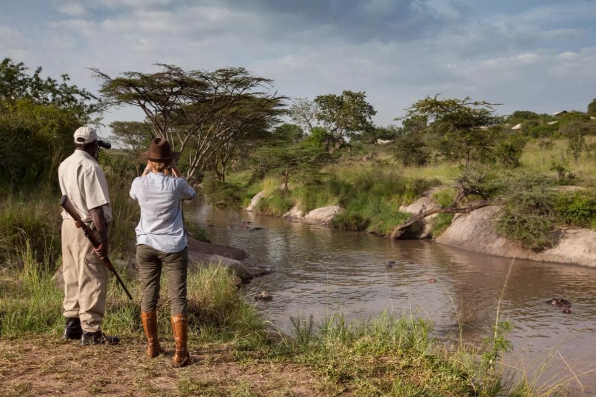 A woman and safari guide watch animals cross a river in the Serengeti. There are several things to have on your Tanzania safari packing list