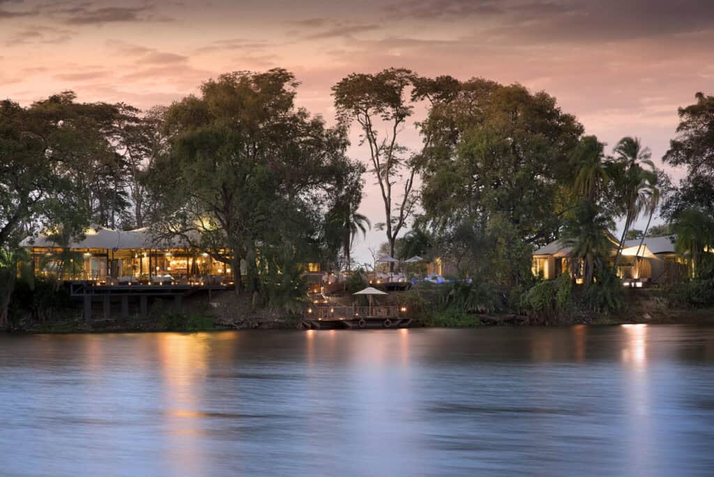 A lodge at dusk with lights and trees overlooking the Zambezi River